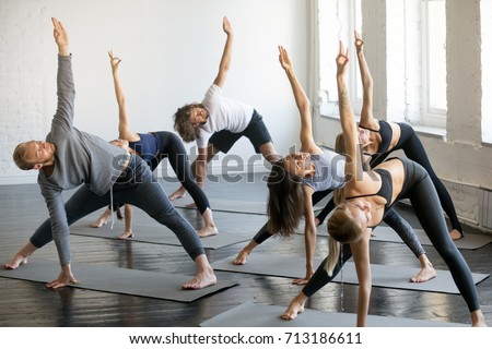 Group of young sporty people practicing yoga lesson with instructor, standing in Trikonasana exercise, extended triangle pose, working out indoor, full length studio. Wellbeing, wellness concept  Royalty-Free Stock Photo #713186611