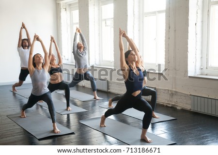 Group of young sporty attractive people practicing yoga lesson with instructor, standing together in Virabhadrasana 1 exercise, Warrior one pose, working out, indoor full length, studio background  Royalty-Free Stock Photo #713186671