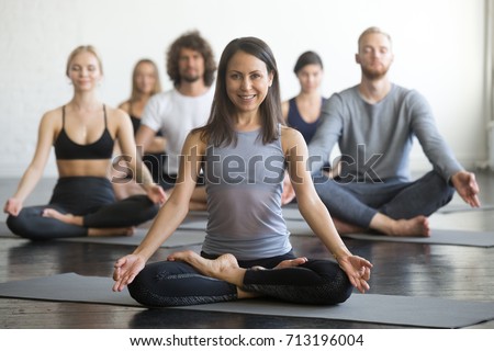 Group of young sporty people practicing yoga lesson with instructor, sitting in Sukhasana exercise, Easy Seat pose, friends working out in club, focus on woman in Padmasana, indoor full length, studio Royalty-Free Stock Photo #713196004