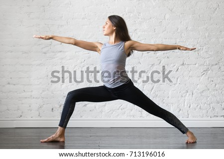 Young attractive woman practicing yoga, standing in Warrior one exercise, Virabhadrasana II pose, working out, wearing sportswear, gray tank top, black pants, indoor full length, studio background  Royalty-Free Stock Photo #713196016