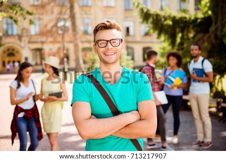 Close up focused photo of young successful blond nerdy student, standing with crossed arms and smiles, behind are his classmates, park near college, sunny day, carefree and enjoyable mood