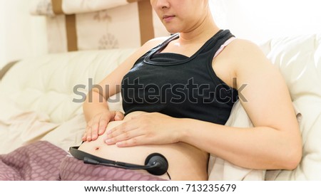 Smile Asia white woman of 33 weeks Pregnancy holding headphones on belly for baby to listen sitting on bed
