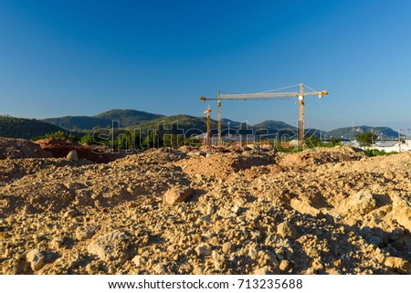 Ground level of new building construction site near the mountain with many yellow cranes against blue sky.