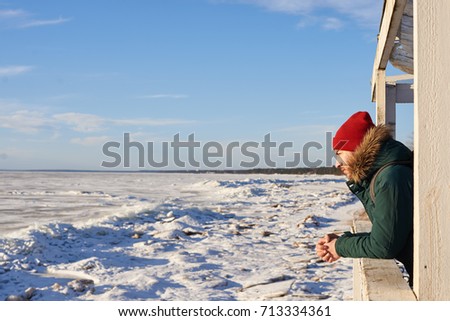 Lifestyle picture of attractive young Caucasian male in coat and red hat standing on porch of house, enjoying warm winter day and beautiful snowy landscape with nobody around. People and nature.