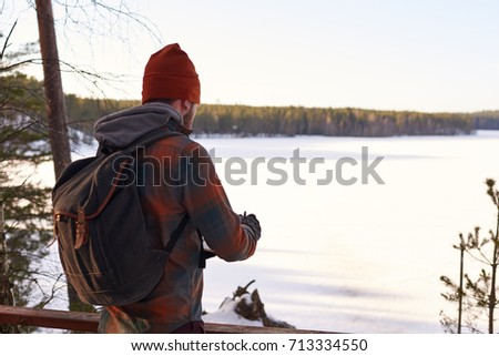 Wintertime, wild nature, traveling and active listyle concept. Back view of bearded male backpacker standing in front of vast fields covered with snow and ice, writing thoughts in his copybook.