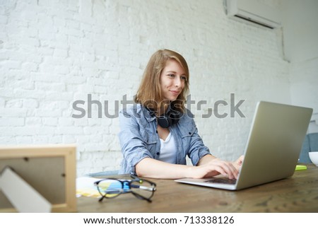 Indoor shot of positive self-employed young female keyboarding on generic laptop pc while working in her office, sitting at white brick wall. Cheerful student girl using wifi on notebook computer.