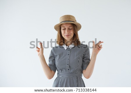 Picture of pretty girl in romantic dress and hat having excited, superstitious and naive look, keeping fingers crossed and eyes closed for good luck, hoping her dreams will come true. Body language.