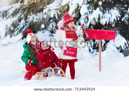 Happy children in knitted reindeer hat and scarf holding letter to Santa with Christmas presents wish list at red mail box in snow under Xmas tree in winter forest. Kids sending post to North Pole