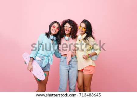 Graceful girl in yellow shirt with leather backpack posing near african curly friend in jeans. Cheerful black young woman in sunglasses standing between european and latino ladies on pink background.