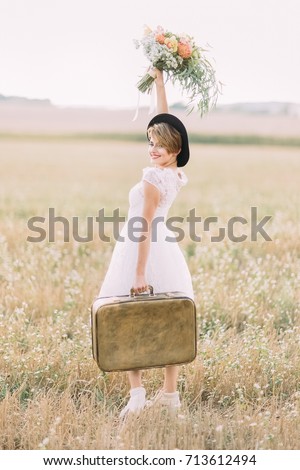 The side view of the smiling bride carrying the vintage suitcase, keeping up the colourful bouquet and looking at the camera at the background of the sunny field.
