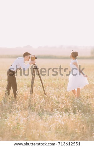 The side portrait of the vintage dressed newlyweds in the field. The groom is taking photos while the bride is posing.