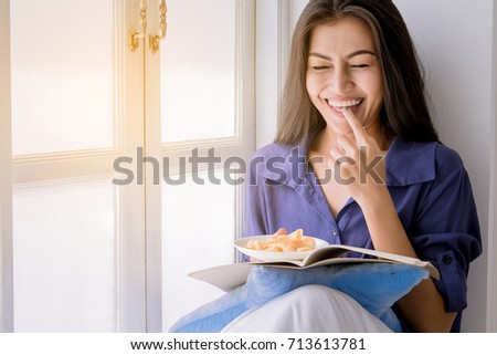 happiness woman enjoy eating chips while reading a book beside window at home in the morning. chill out concept
