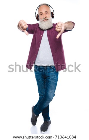 senior bearded man showing thumbs down while listening music with headphones, isolated on white