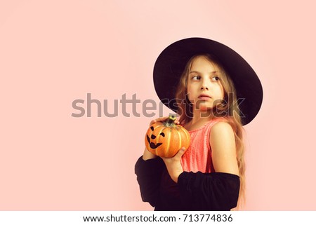 Child in witch costume and jack o lantern. Girl with carved orange pumpkin isolated on pink background, copy space. Kid in black witch hat, dress and tricky face. Halloween and autumn concept