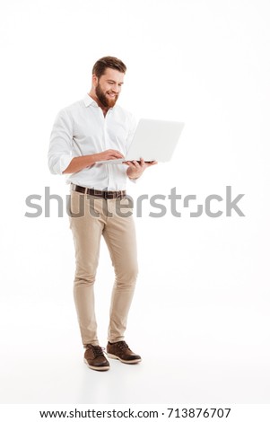 Picture of handsome young bearded man standing over white wall background isolated. Looking aside using laptop.