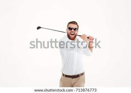 Picture of handsome young screaming emotional bearded man standing over white wall background isolated holding golfstick. Looking at camera.
