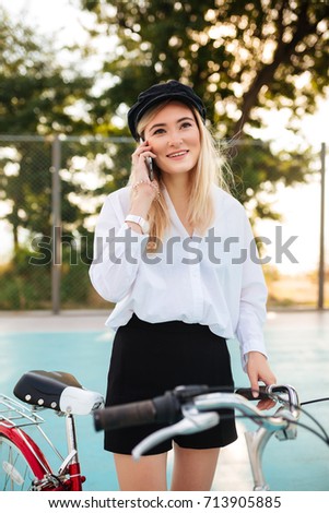 Young pretty girl with blond hair in shirt and shorts standing with red bicycle on basketball court in park and talking on her cellphone. Portrait of smiling lady in black peaked cap looking aside 