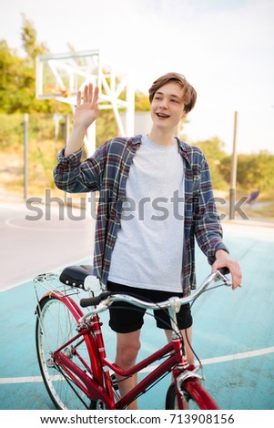 Young man with blond hair in shorts and casual shirt waving and showing hello gesture while standing with bicycle on basketball court. Portrait of smiling boy holding bicycle and happily looking aside