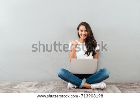Happy smiling asian woman working on laptop computer while sitting on the floor with legs crossed and pointing finger away isolated over gray background Royalty-Free Stock Photo #713908198