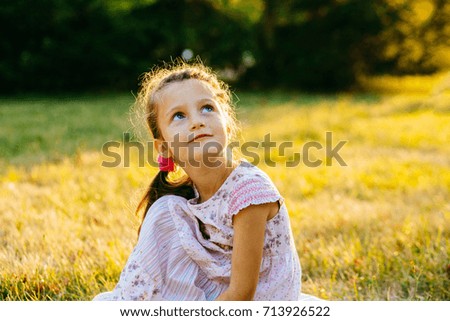 Cute preschooler girl with blue eyes sitting on the grass in sunset in summer time.