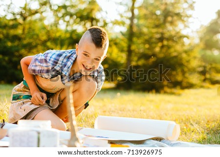 Cute European boy laughing and looking at camera in summer sunny day. Teenager boy drawing picture outdoor in park. Art, drawing and kids creativity concept.