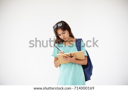 People, youth, education, learning and knowlegde concept. Picture of beautiful college student girl wth glasses on head carrying backpack and writing down in copybook, posing isolated in studio