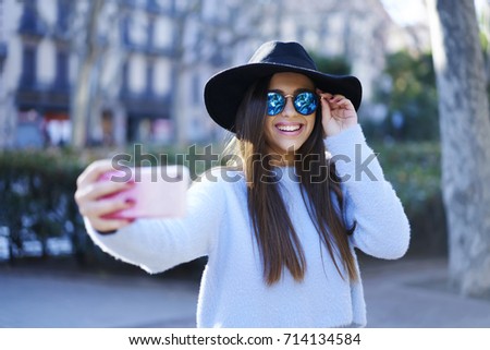 Attractive smiling hipster girl dressed in stylish clothing streaming online video for friends in social networks on smartphone.Pretty cheerful traveler taking self portrait while walking outdoors
