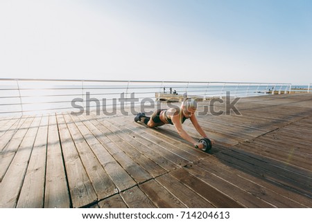 The young beautiful athletic girl with long blond hair in headphones, listening to music and training with dumbbells at sunrise over the sea