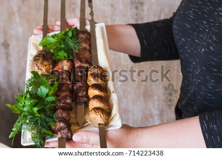 Waiter brings shish-kebab with grilled vegetables on white dish. Meat, field mushrooms and fresh green herbs, barbecue and natural food preparing in restaurant, close up