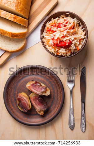 Tasty baked potatoes with slices of bacon in brown clay plate and sauerkraut in clay bowl on wooden background. Slices of smoked bacon on potatoes. Slices of white bread on cutting board.
