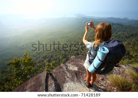 Young woman with backpack standing on cliff's edge and taking a photo
