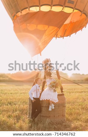 The lovely portrait of the newlyweds in the field. The bride with the wedding bouquet is sitting on the basket of the airballoon while the groom is standing near her.