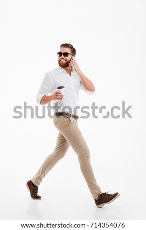 Picture of handsome young bearded man walking over white wall background isolated drinking coffee. Looking aside talking by phone.