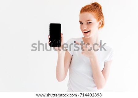 Happy ginger woman showing blank smartphone screen and pointing at him over white background