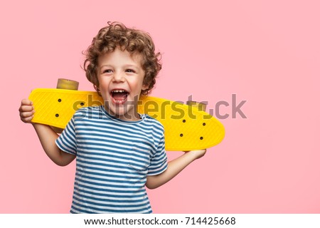 Charming curly boy holding yellow longboard and looking away on pink background.  Royalty-Free Stock Photo #714425668