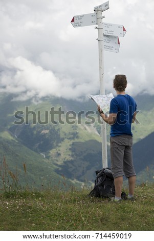 A hiker with a frizzy hair in a blue top standing near the information pole in the Caucasus mountains holding paper map in his hands. Deep valley and slopes partly covered by clouds in the background.