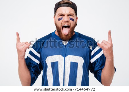 Picture of emotional man fan in blue t-shirt standing isolated over white background. Looking camera showing tongue and rock gesture.