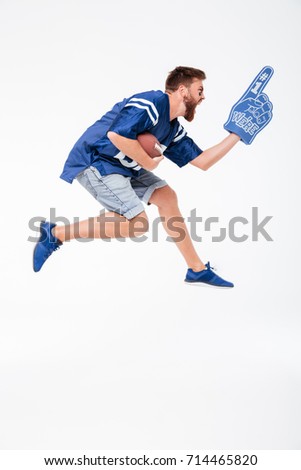 Picture of screaming man fan in blue t-shirt jumping isolated over white background. Looking aside holding rugby ball.