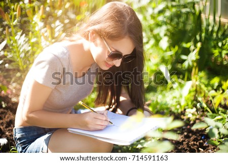 Attractive young artist works on plain air, drowing in pencil. Wearing sunglasses, smiling. Enjoying painting on summer day. Art education, art college.
