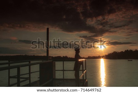 Woman with bicycle helmet standing on a bridge at a lake at sunset. The setting sun is reflected in the water of the lake. In the sky there are sunset-lighted clouds.