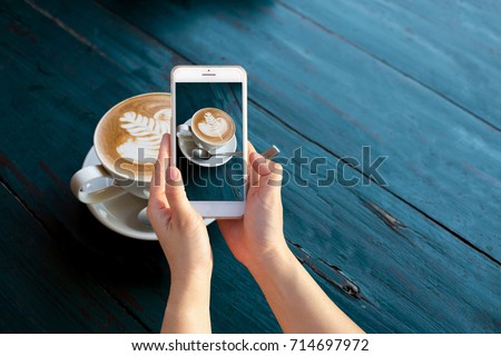 Hand holding mobile smart phone or cell telephone digital camera taking photo to Cup of Hot Cappuccino coffee with fresh milk Art's surface floating on top for Post upload file to online on Internet .