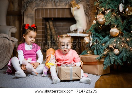 Two little sisters sitting and playing, smiling near Christmas tree, box presents, fireplace on white wooden floor indoor