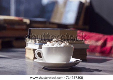 A white ceramic cup stands on the table in the background of books. A cup of coffee with foam and chocolate. A cafe