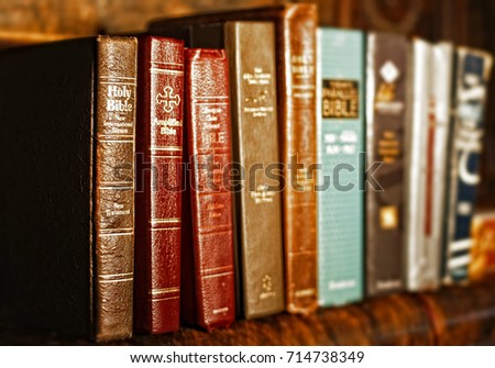 Numerous Holy Bibles sit in a row on an antique wood shelf.  The well-used bibles are various translations, versions, and ages.  Shallow depth of field, with focus point on the far left bibles.