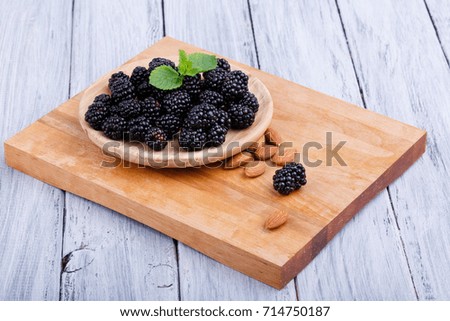 A top view of a beautiful composition of a plate with a heap of juicy and raw blackberries on a gray wooden background. Healthy, fresh and ripe berries, mint and spicy almonds on a cutting board.