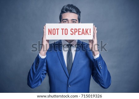 Young attractive man in a blue suit holding a white sign with red inscription  NOT TO SPEAK on a gray background. Toned