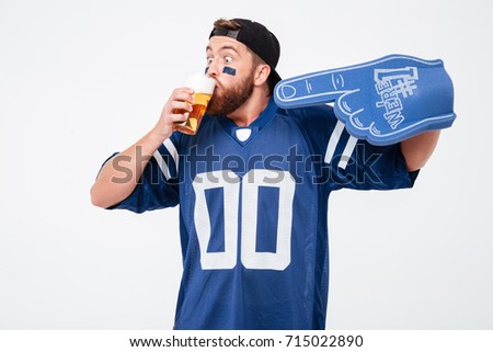 Picture of emotional man fan in blue t-shirt wearing fan finger number one glove standing isolated over white background. Looking aside drinking beer.