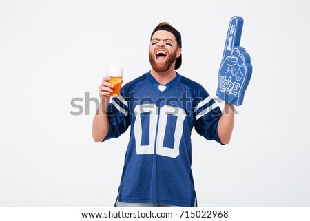 Picture of emotional man fan in blue t-shirt wearing fan finger number one glove standing isolated over white background. Looking aside drinking beer.