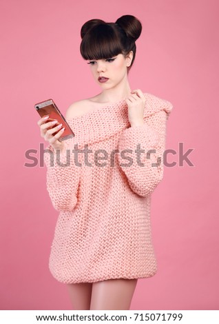 Brunette holding cell telephone. Fashion studio teen look style over pink. Fashionable young girl wears wool sweater posing isolated on studio background.