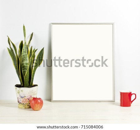 Silver frame mockup with plant pot, mug and apple on wooden shelf. Empty frame mock up for presentation design. Template framing for modern art.